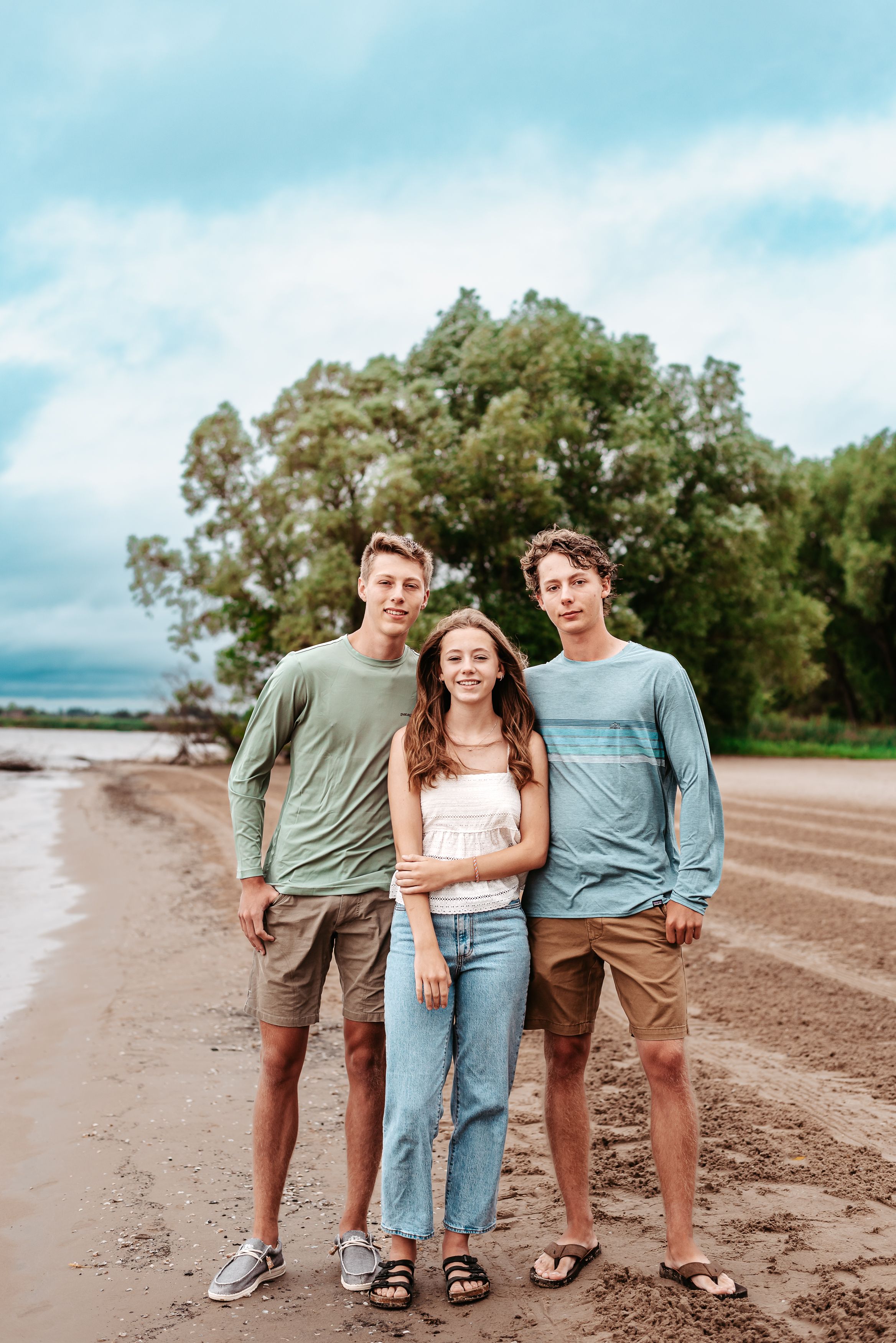 Three people in casual clothing stand together on beach with trees in background.
