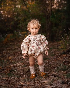 Toddler wearing floral romper and brown shoes stands on forest path.