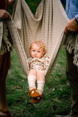 Baby in floral outfit sitting in grey blanket swing held by adults outdoors.