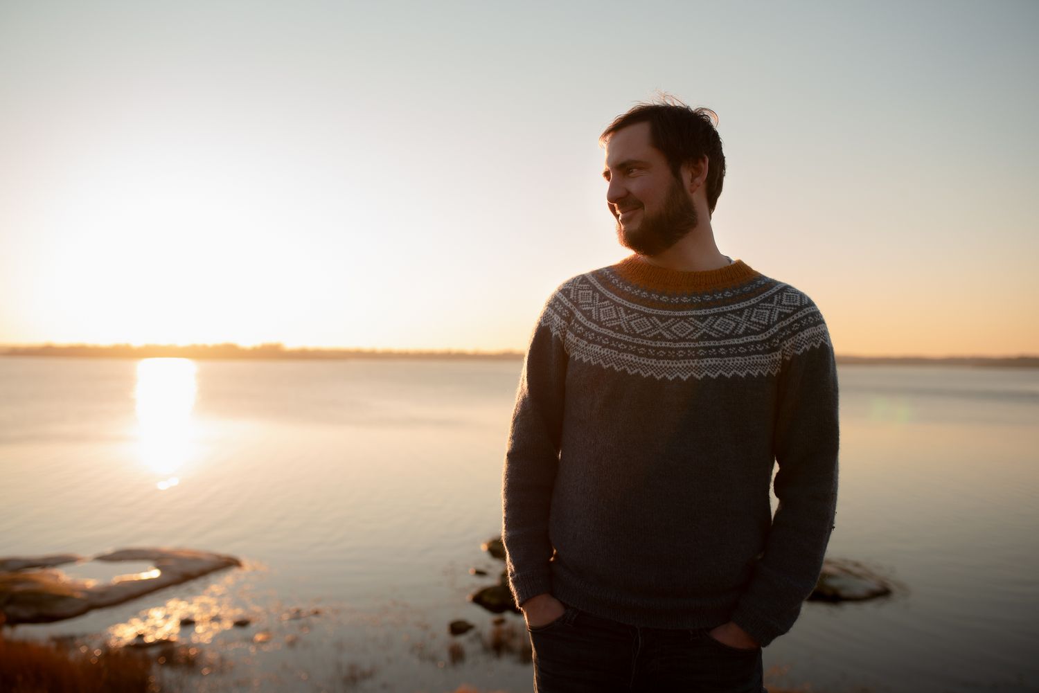 Person in a patterned sweater stands contemplatively by the water at sunset.