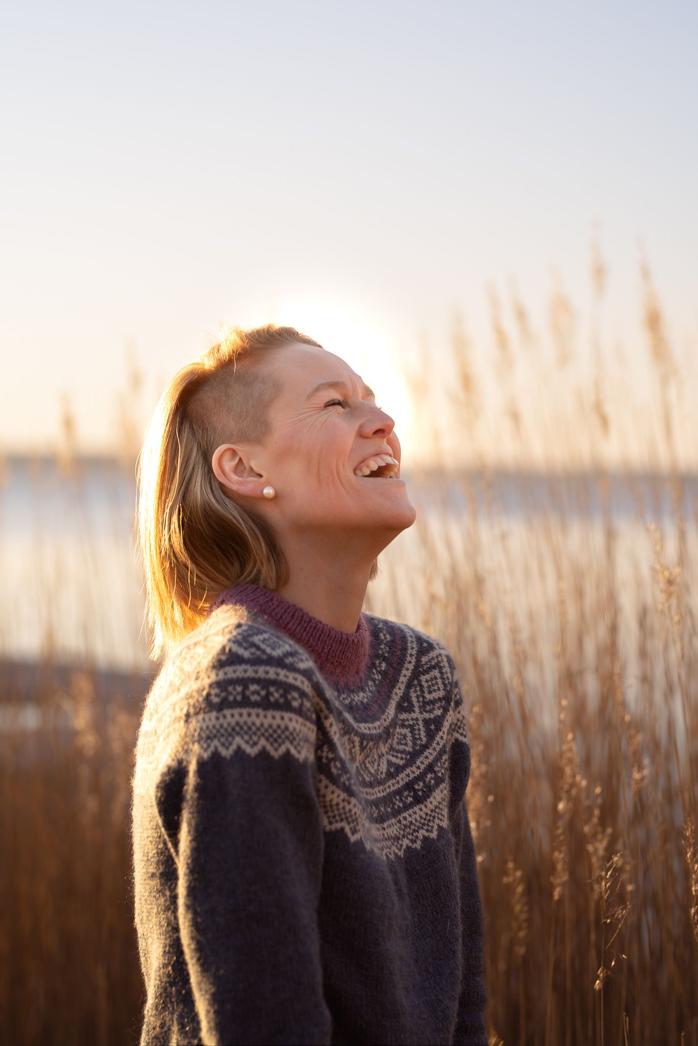 Person in patterned sweater laughing joyfully against golden sunset and tall reeds.