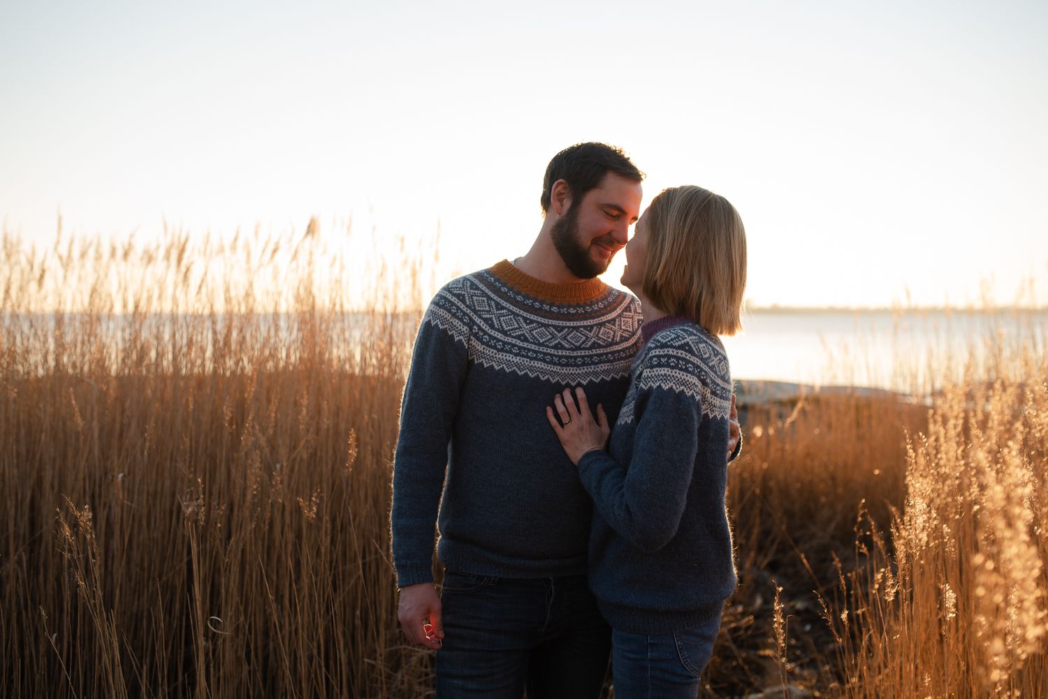 A couple wearing matching patterned sweaters embrace in a field of tall grass at sunset.