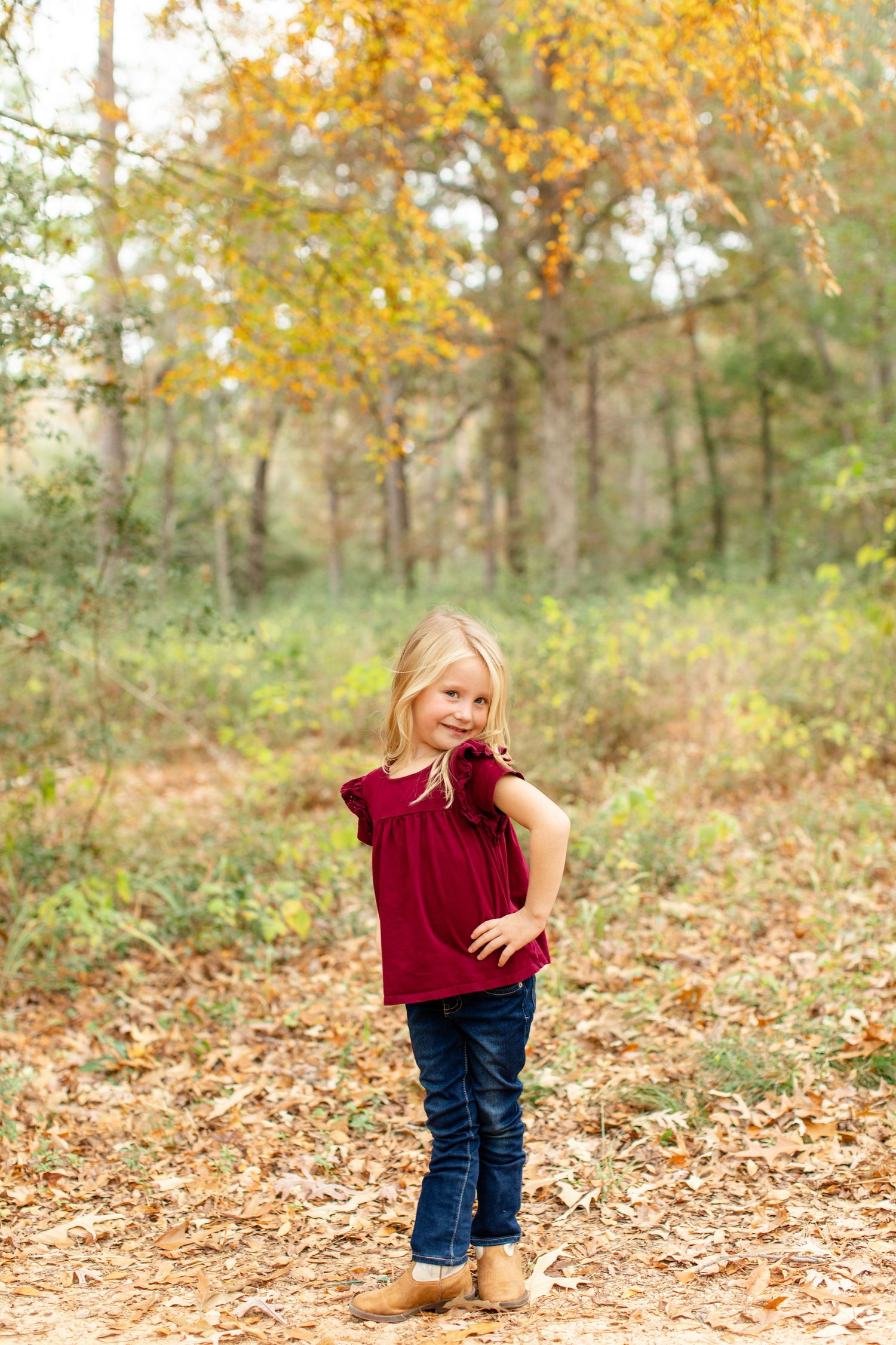 Small child in red top striking pose on forest path covered in fall leaves.