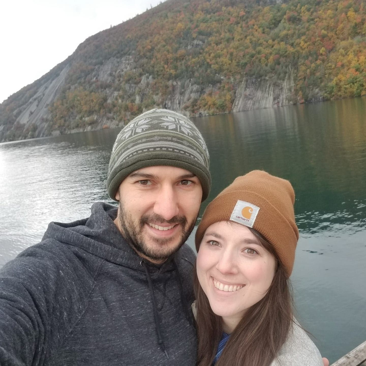 A couple takes a selfie against a scenic mountain lake backdrop wearing winter hats.