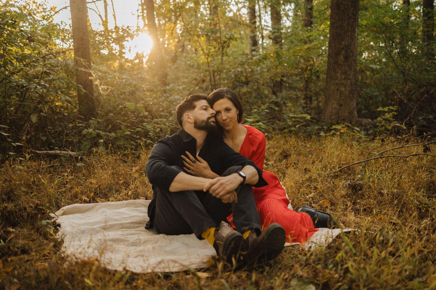 Romantic couple sitting on blanket in autumn forest at sunset with golden light filtering through trees.