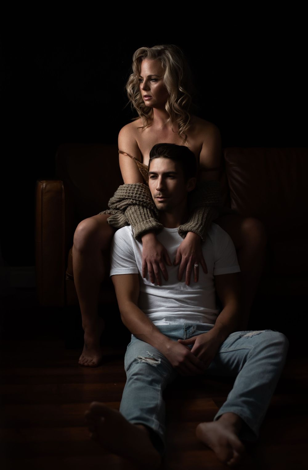 Dramatic moody portrait of a couple in intimate pose on dark leather furniture.