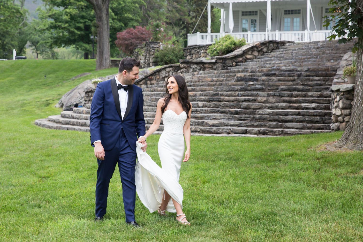 Newlyweds walk together on landscaped grounds with stone steps and mansion in background.