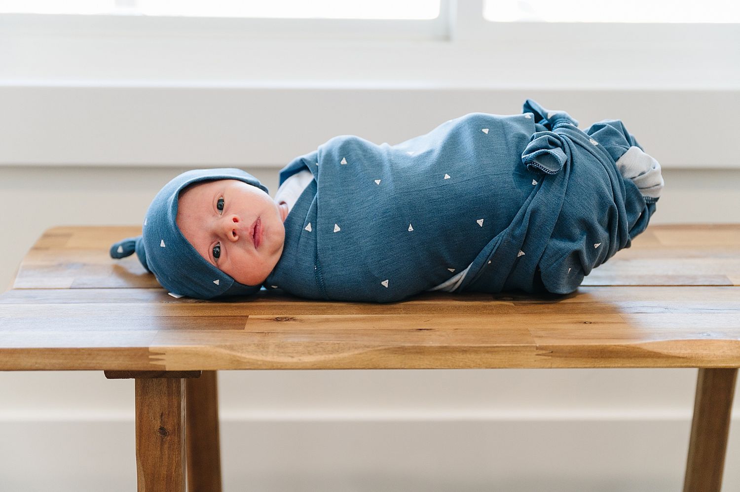 Baby wearing blue polka dot swaddle and hat lying on wooden table.