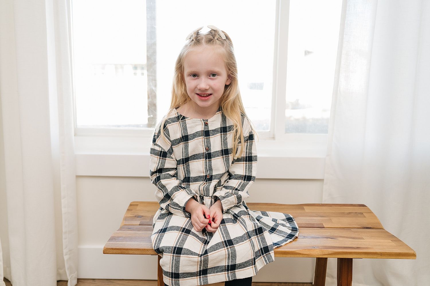 Child in plaid dress sitting on wooden bench by window.