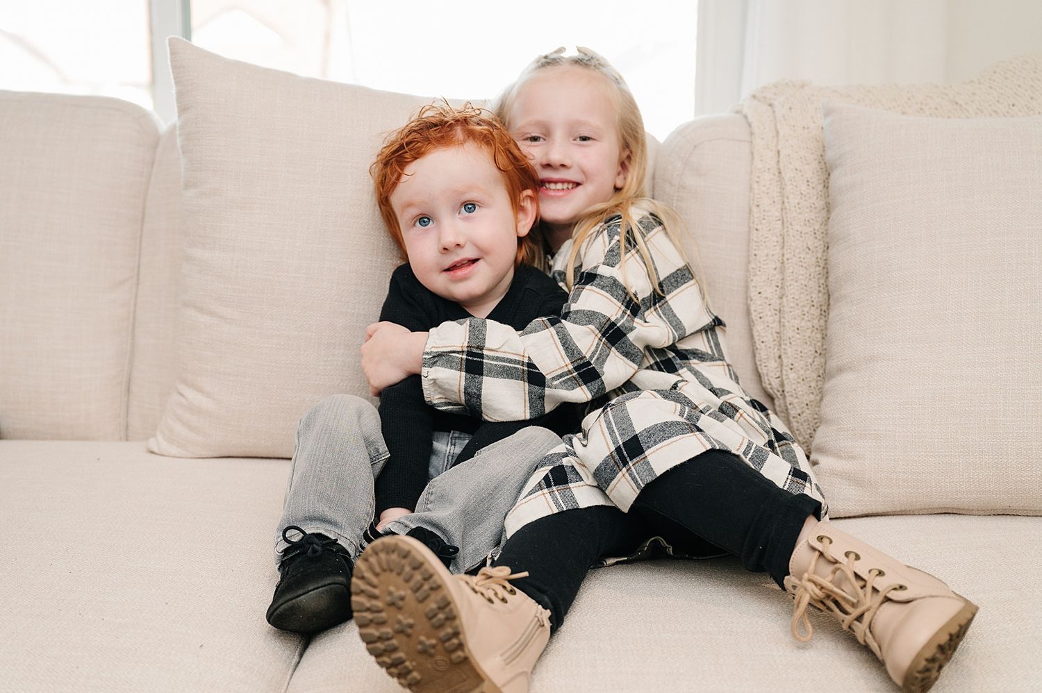 Two children in matching plaid outfits sit together on a beige couch.