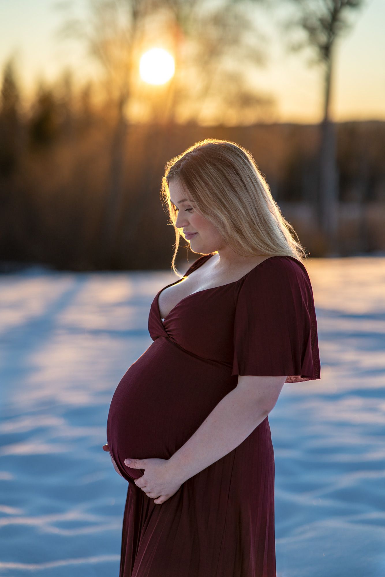 En blond gravid kvinna i vinröd långklänning håller om sin gravidmage i soluppgången. Det är en vintermorgon.
