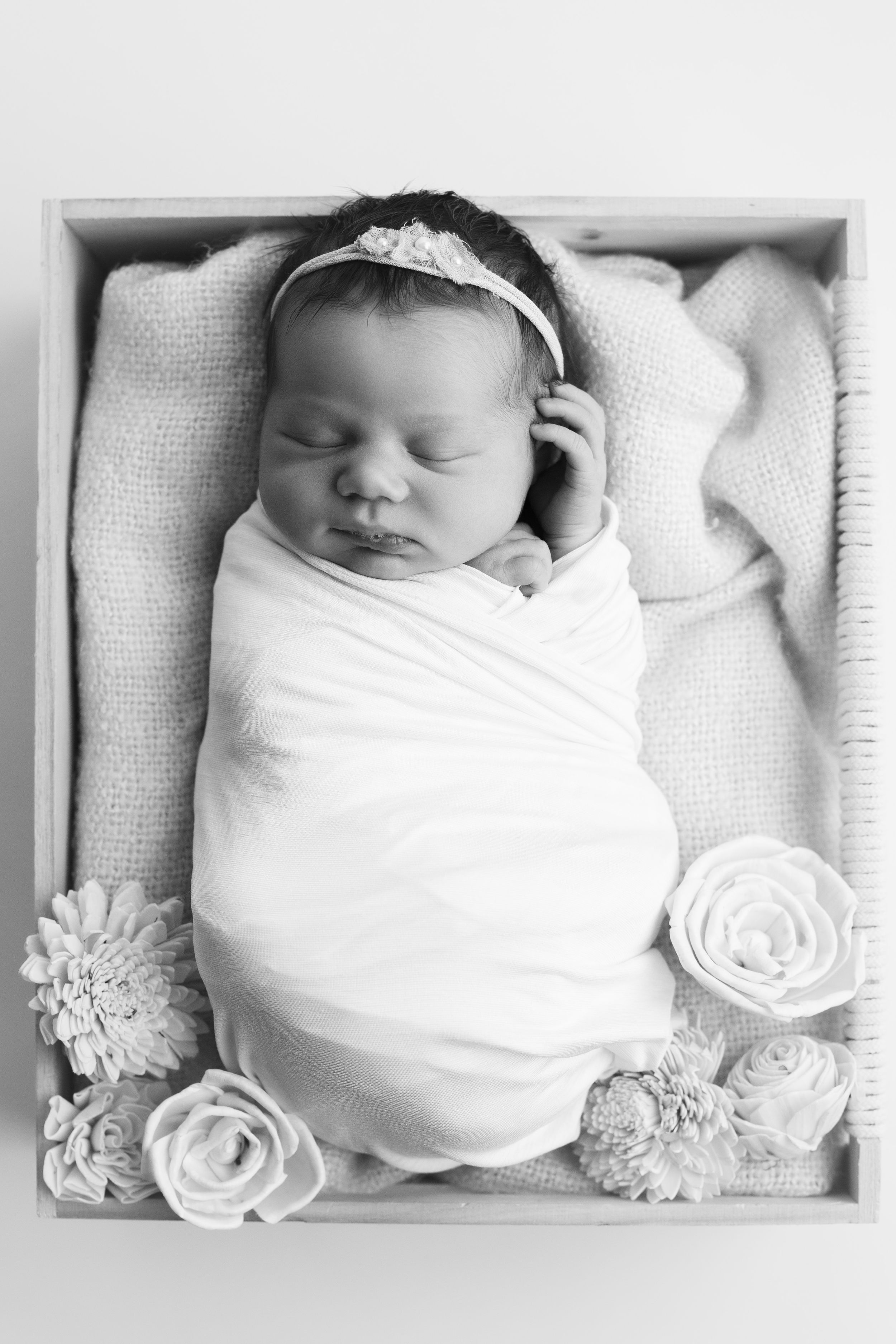 Newborn baby girl in a wooden box, wrapped in a white wrap, and flowers.