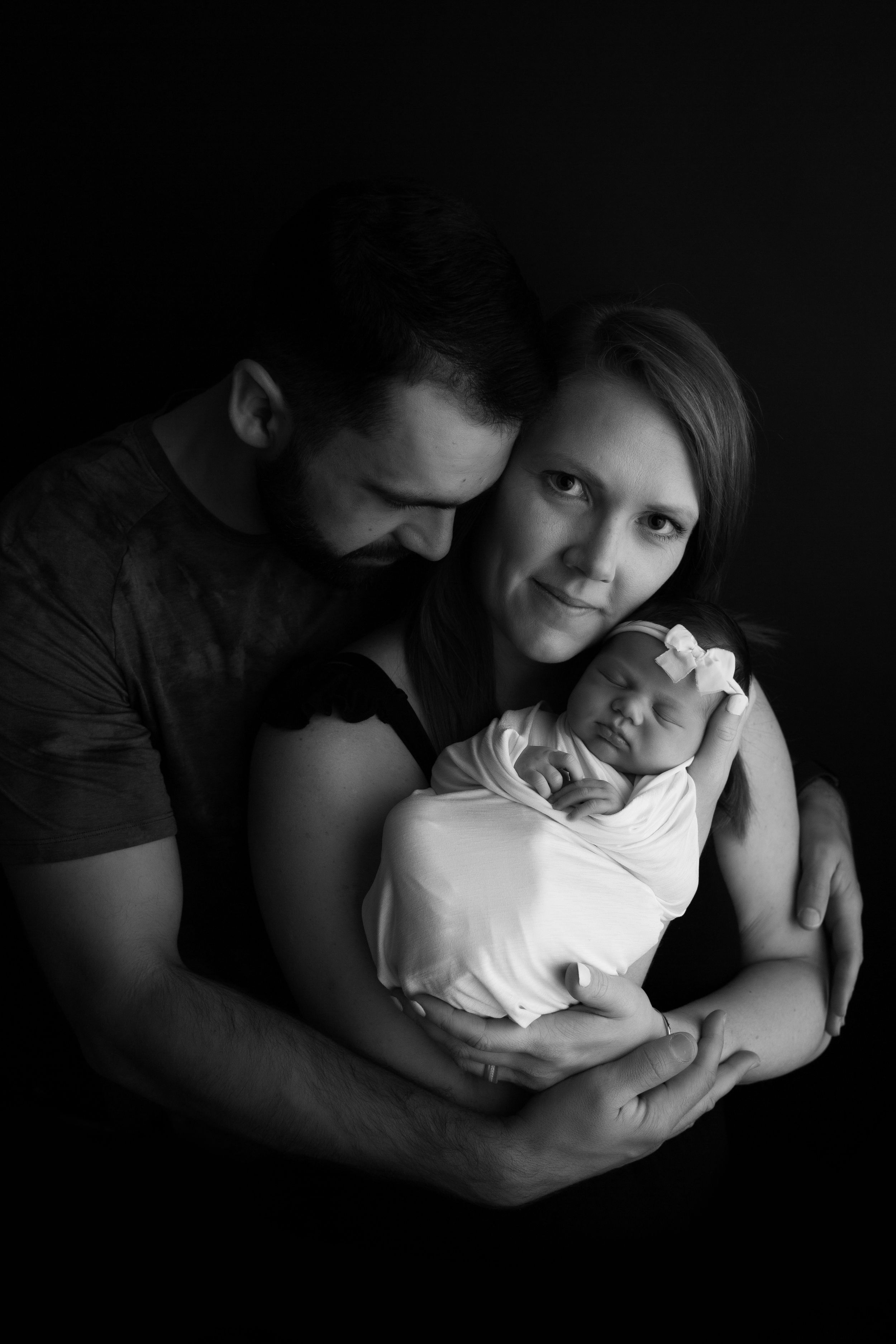 Newborn baby girl cuddled in her mom's arms with dad holding mom on a black backdrop.