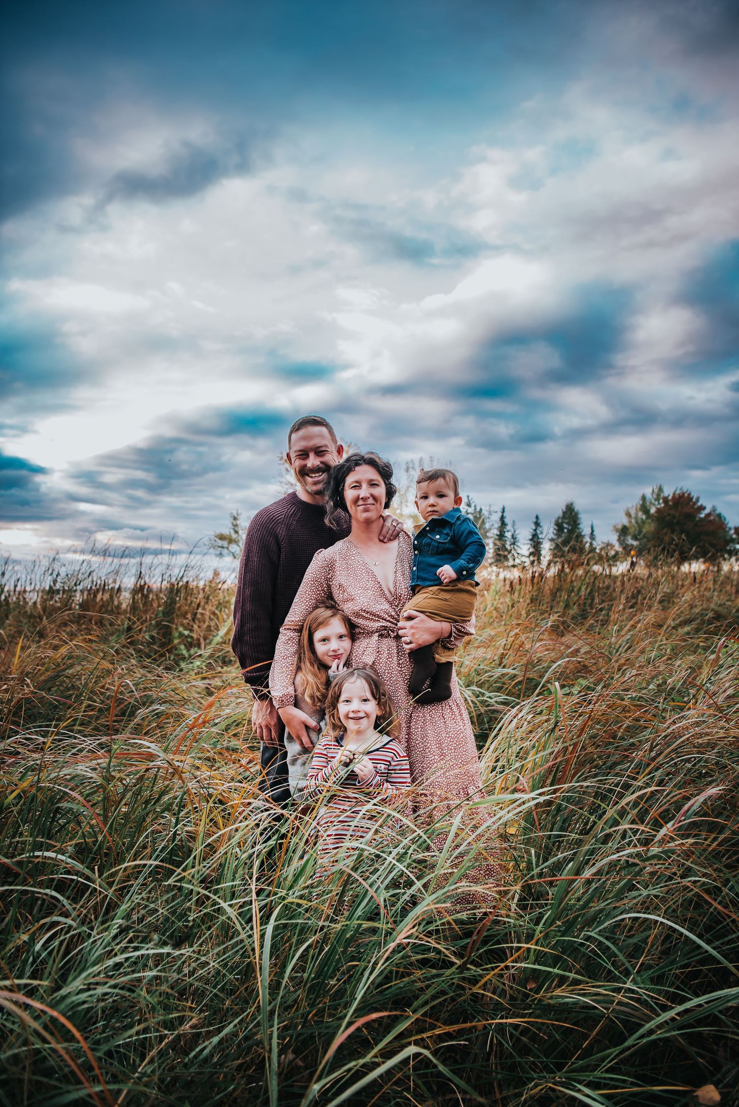 Gorgeous family of 5 stand in the tall grass with a cloudy blue sky behind the.