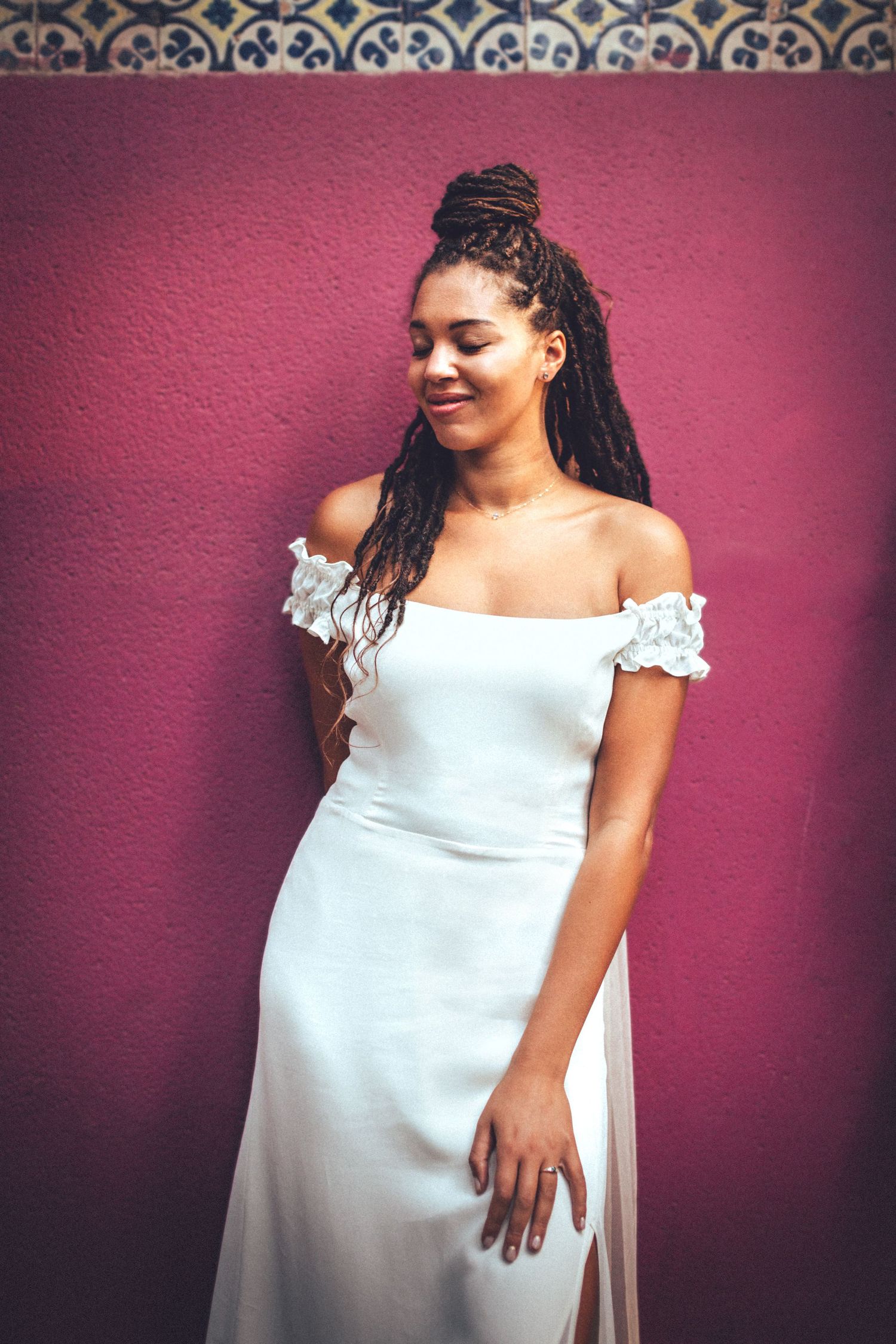 Bride in elegant off-shoulder wedding dress smiling against burgundy wall.