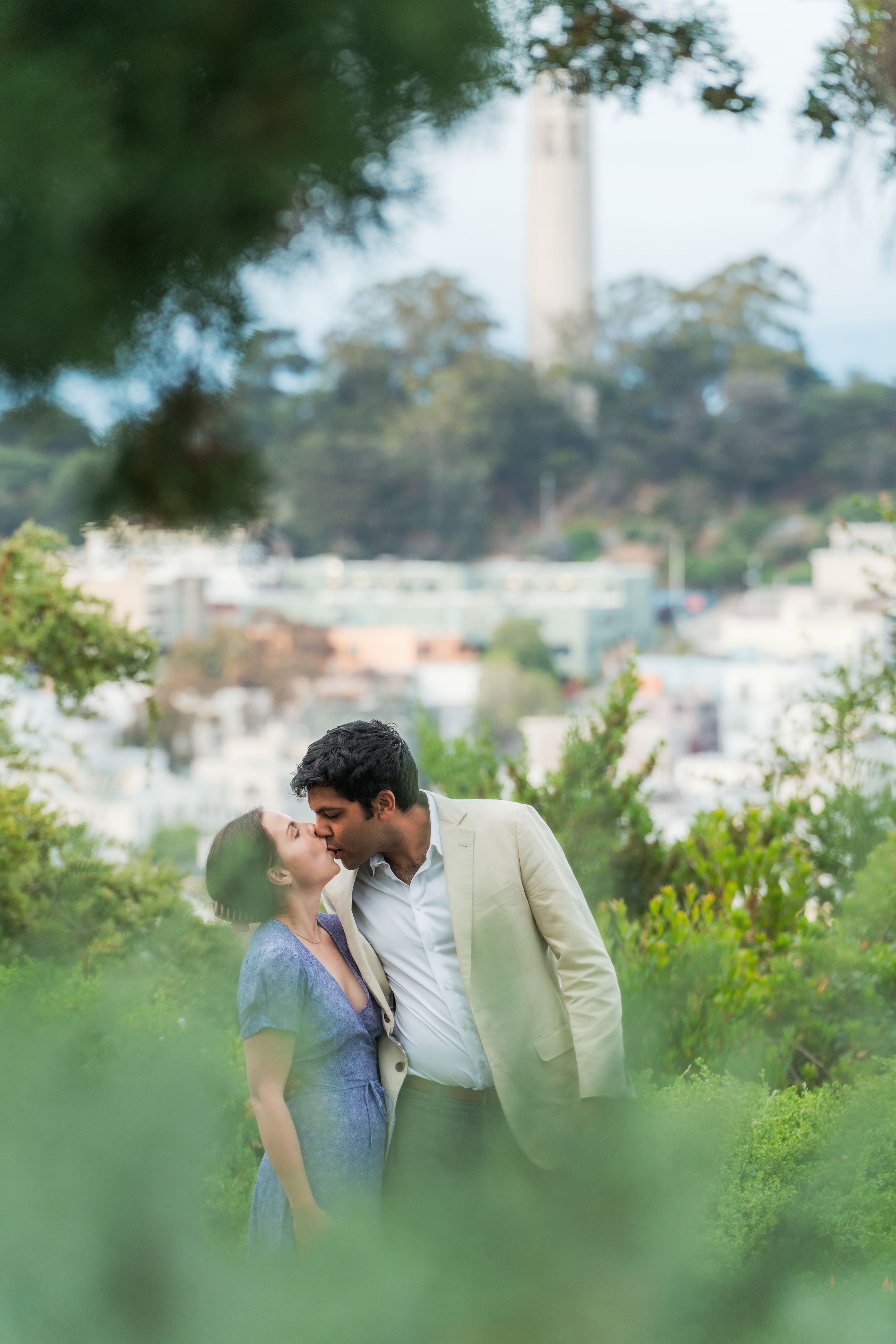 A couple shares a romantic kiss surrounded by lush greenery with Coit Tower visible in the background.