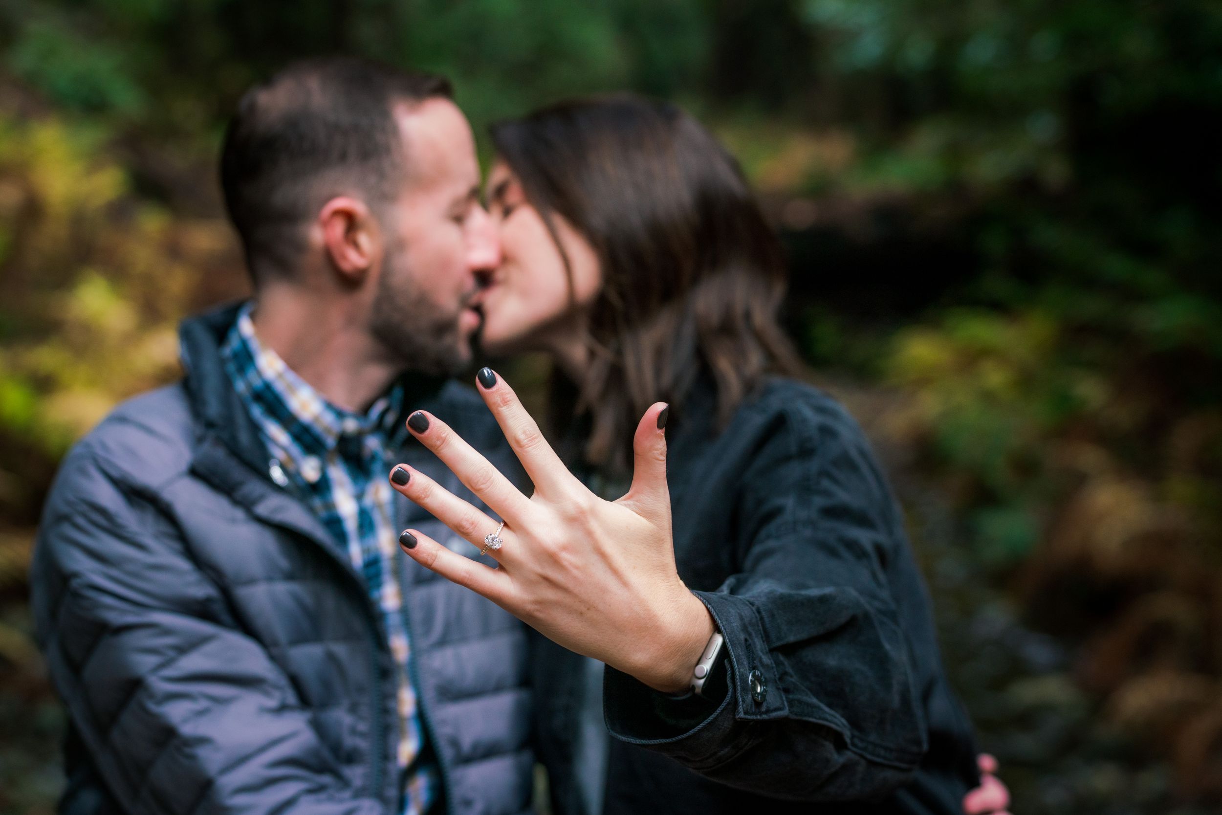 An engaged couple shares a kiss in a forest while the woman displays her engagement ring to the camera.