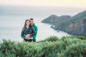 Couple embracing on a coastal hilltop with lush greenery and ocean views at sunset.