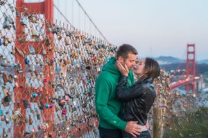 Couple kissing by a love lock fence with the Golden Gate Bridge glowing at sunset behind them.