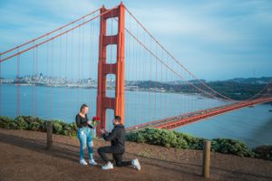 Man proposing on one knee holding roses with the Golden Gate Bridge spanning the bay behind.