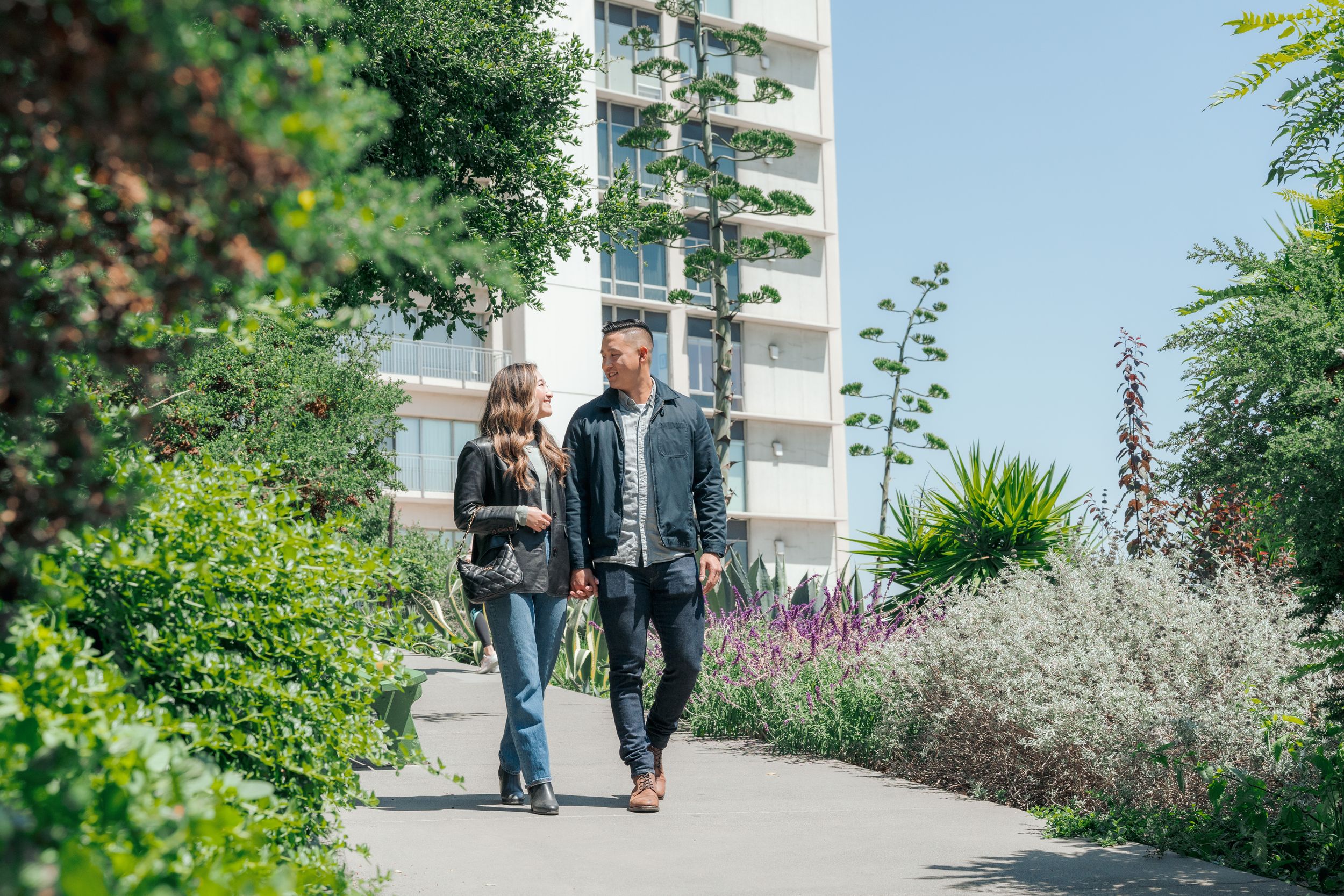 A couple walks hand in hand along a garden path with a modern building in the background.