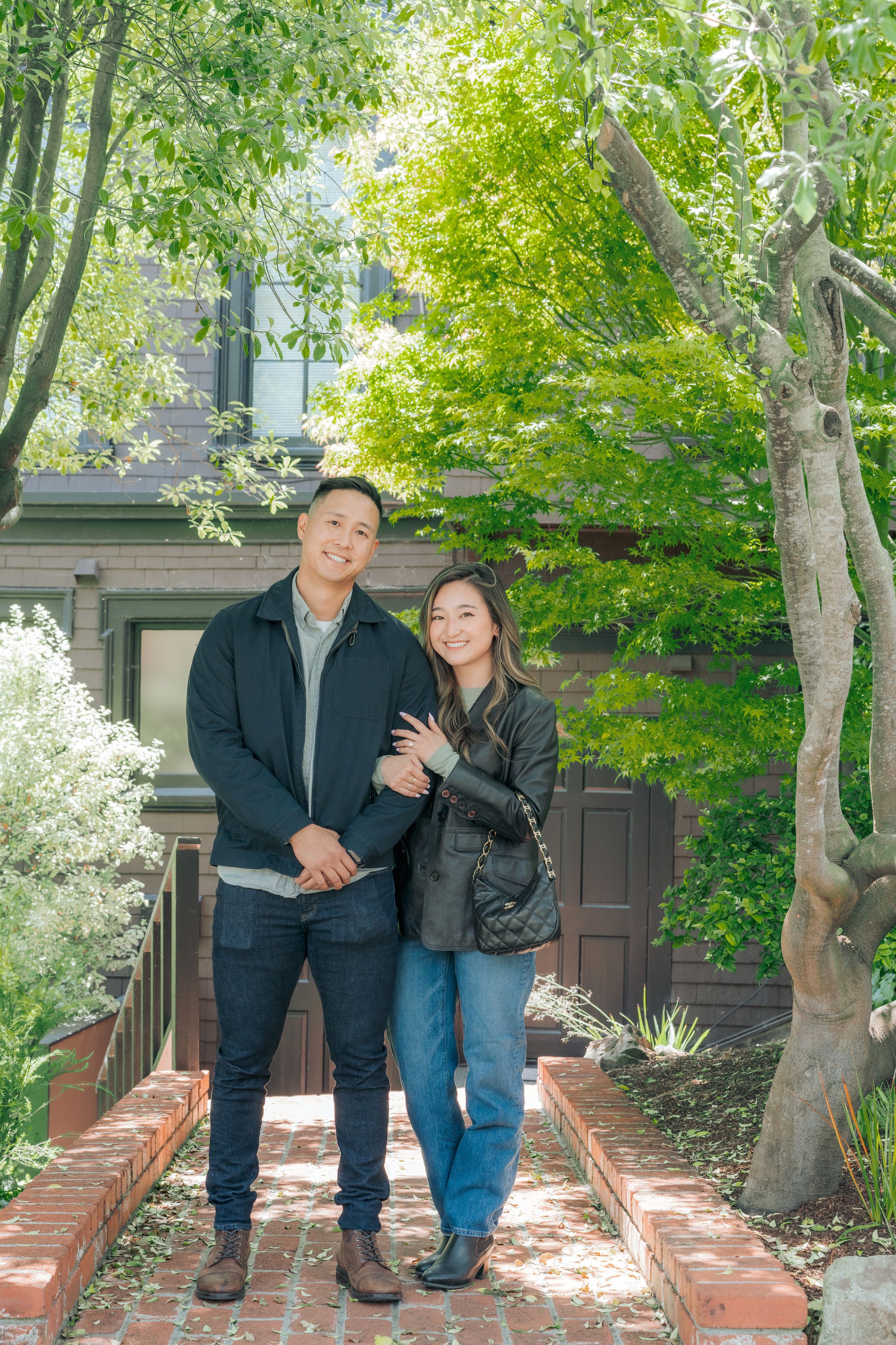 A smiling couple poses together on brick steps surrounded by lush green trees near a home.