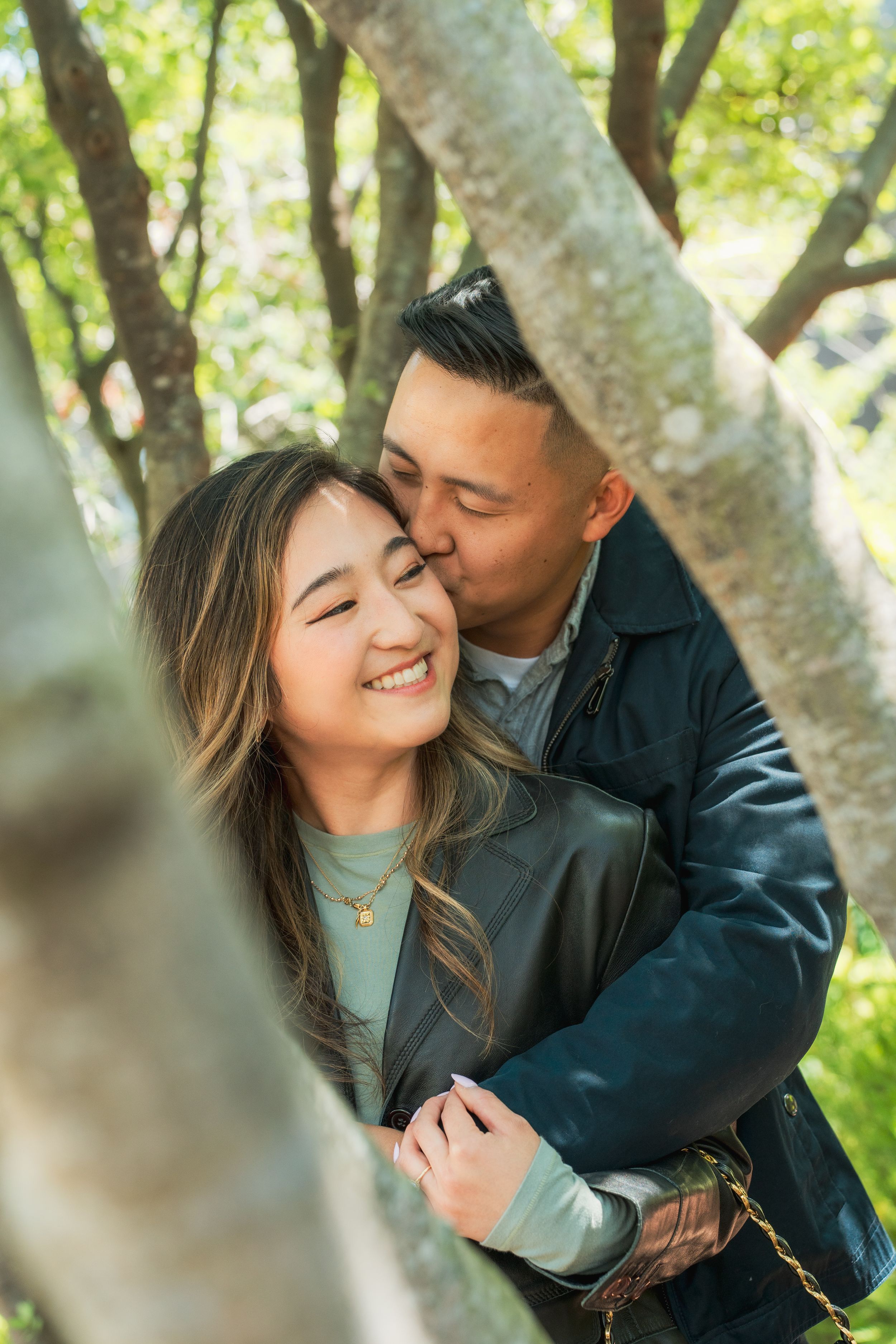 A man tenderly kisses a smiling woman on the cheek while embracing her among sunlit trees.