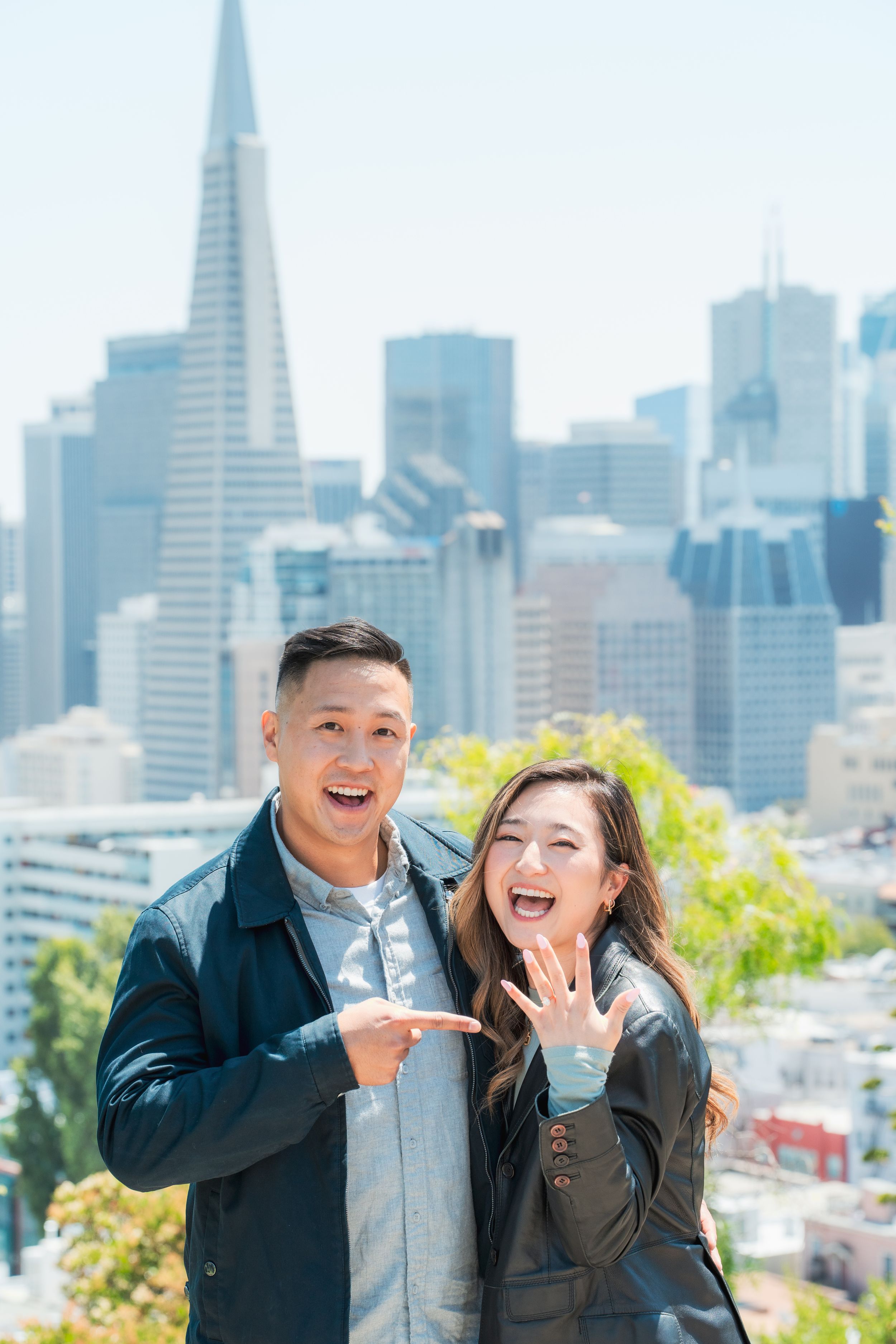 Excited couple showing off engagement ring with San Francisco skyline and Transamerica Pyramid in background.
