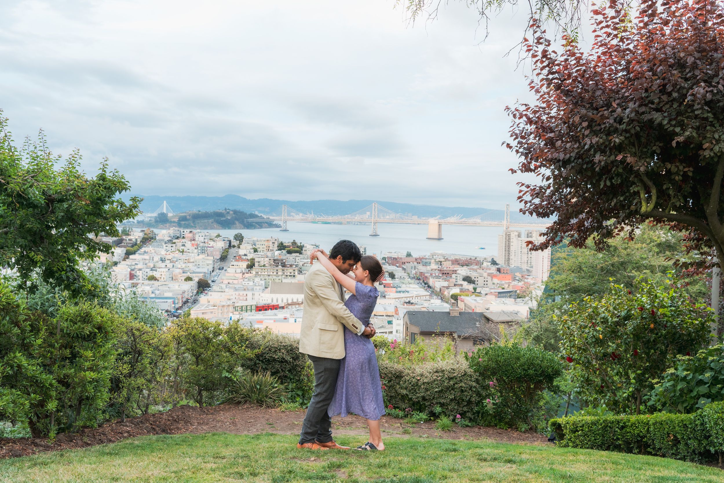 A couple embraces on a grassy hilltop with a panoramic view of San Francisco Bay behind them.