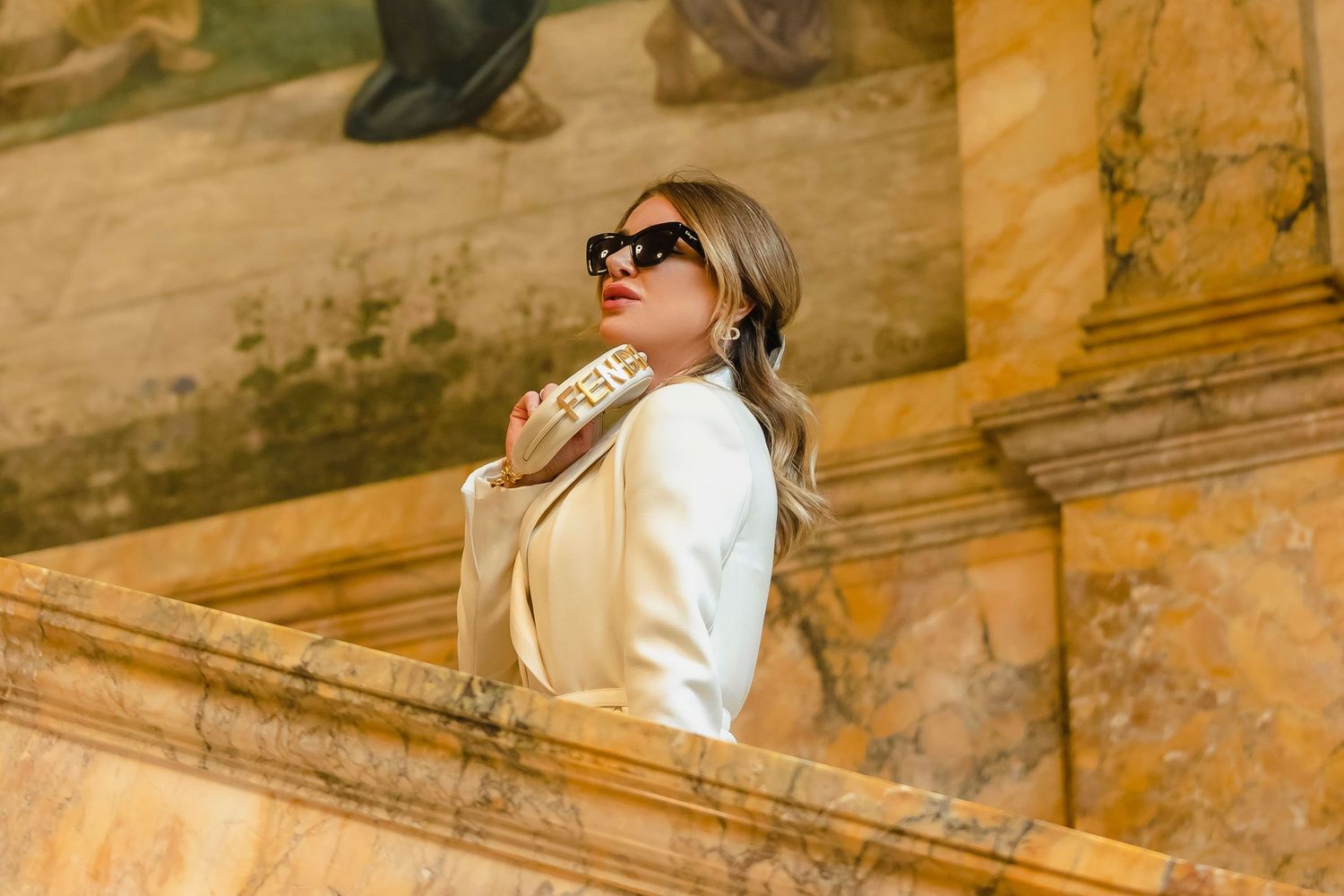 Woman in sunglasses and white sweater sitting on ancient stone steps in warm sunlight at historic architectural site.