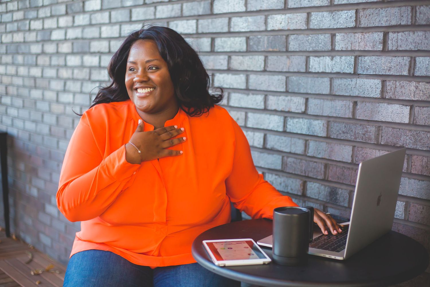 Person in vibrant orange sweater laughs while working on laptop at brick wall cafe.