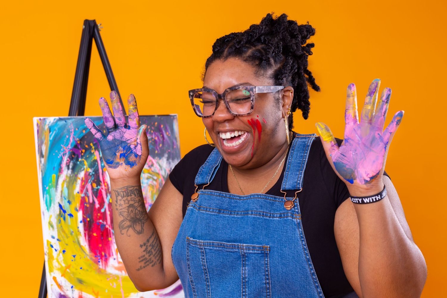Artist with paint-covered hands smiles joyfully in overalls against bright orange background.