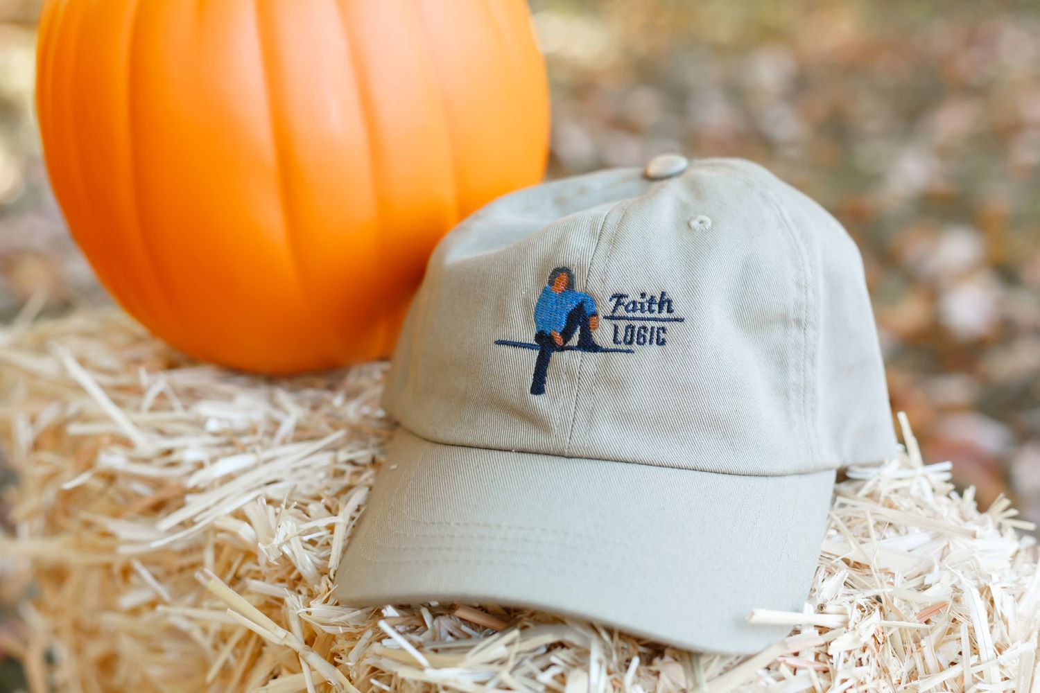 Beige baseball cap with blue bird logo on hay bale next to orange pumpkin.