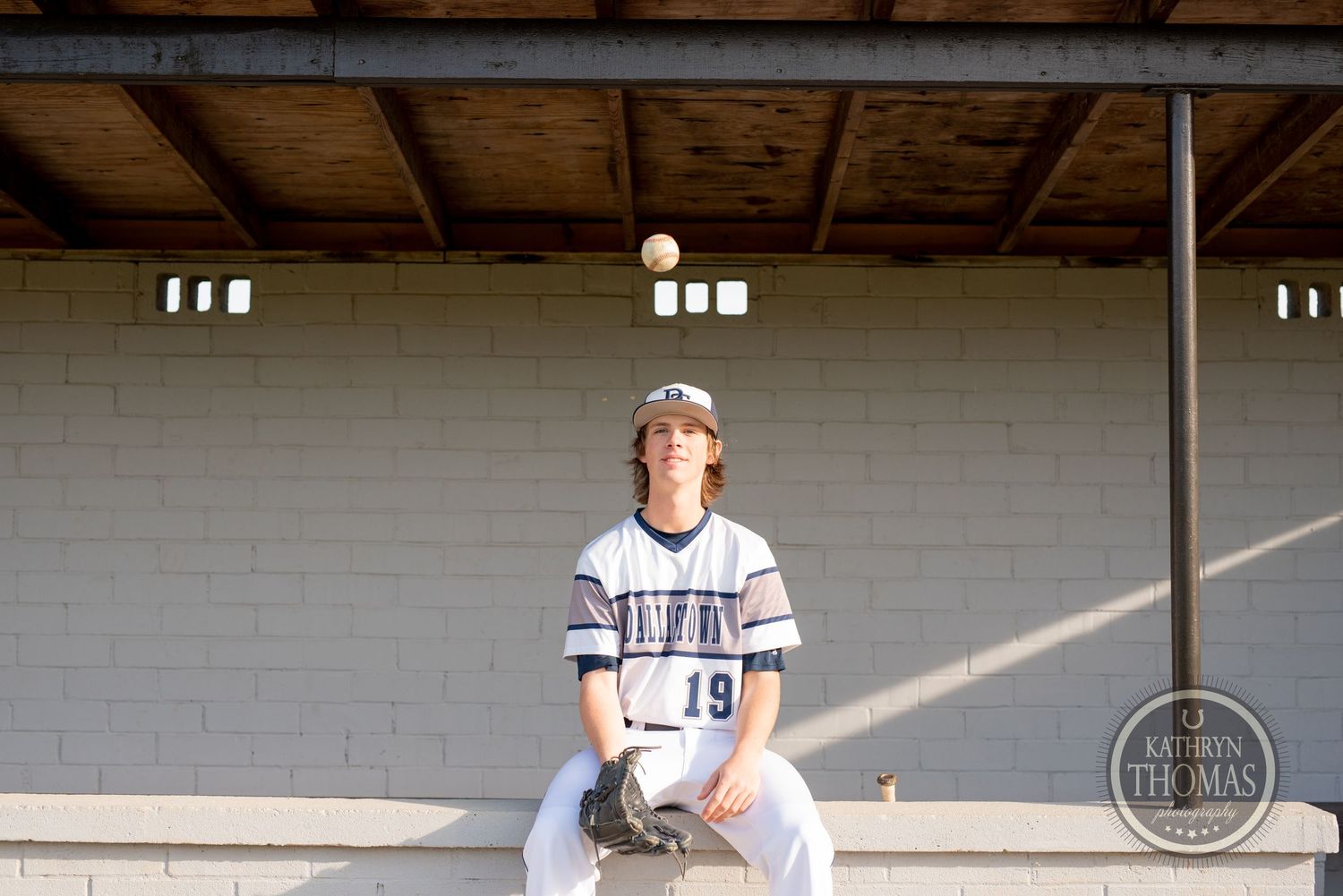 Baseball player sits in dugout with dramatic lighting and white brick wall background.