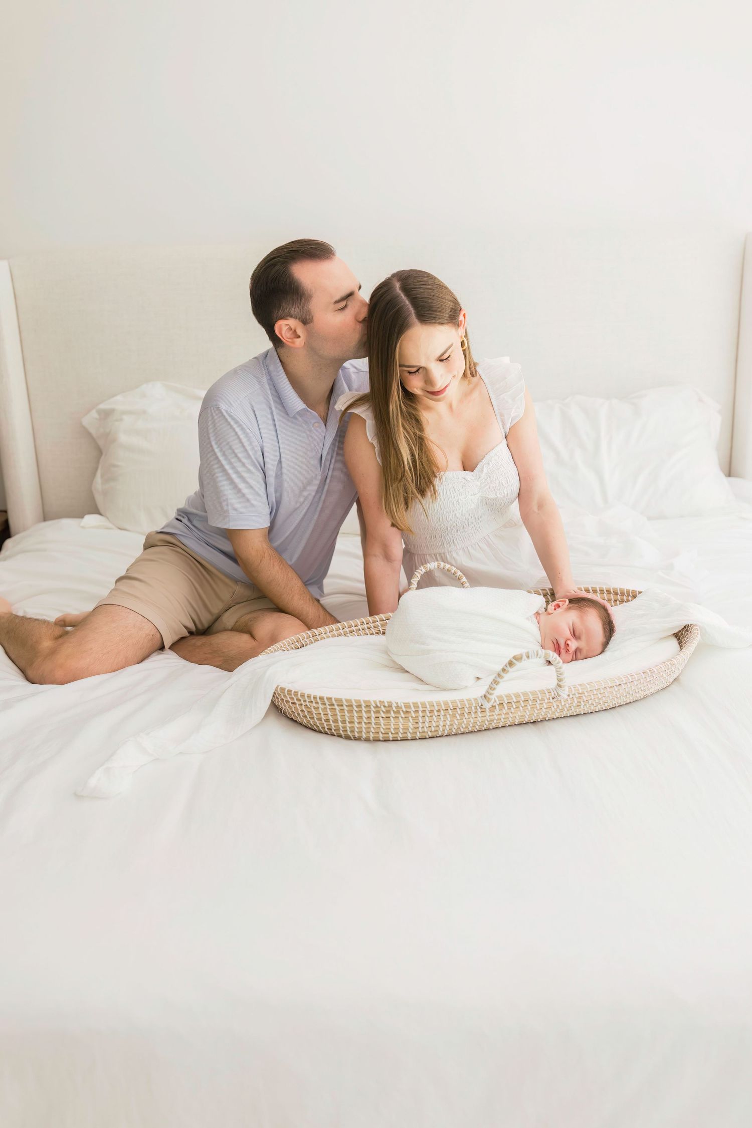 Parents share an intimate moment with their newborn baby resting in a woven bassinet on white bedding.