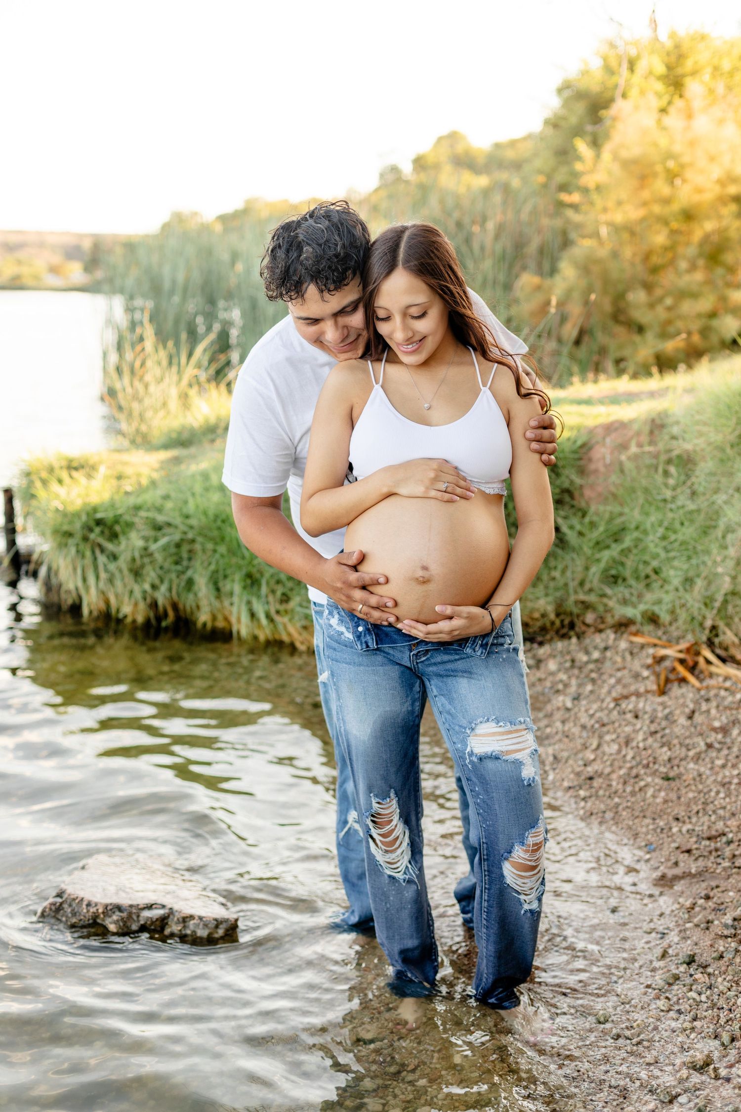 A maternity photo of an expectant couple standing by a lake at sunset.