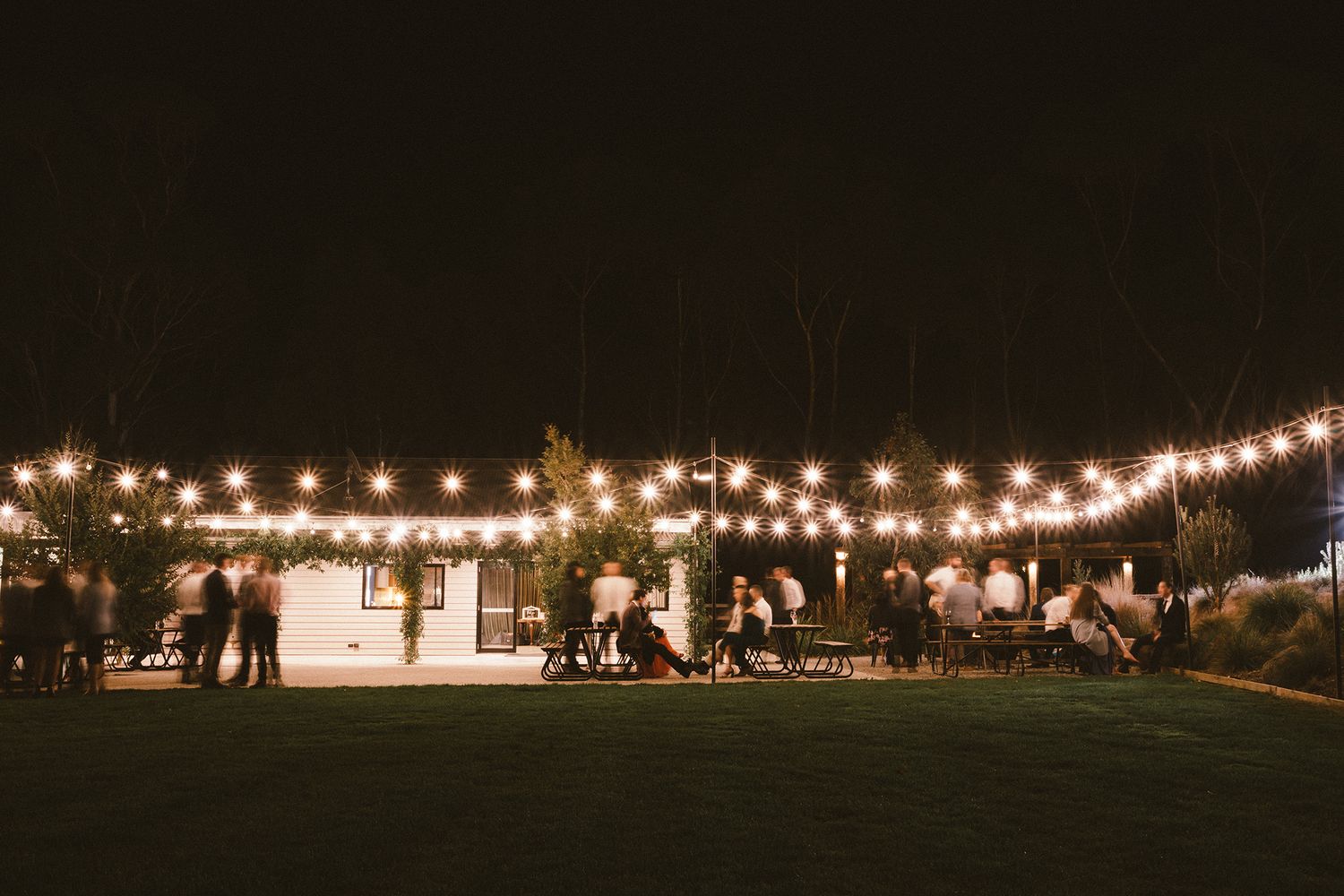 String lights illuminate an outdoor evening reception area with guests gathered on a grassy lawn.