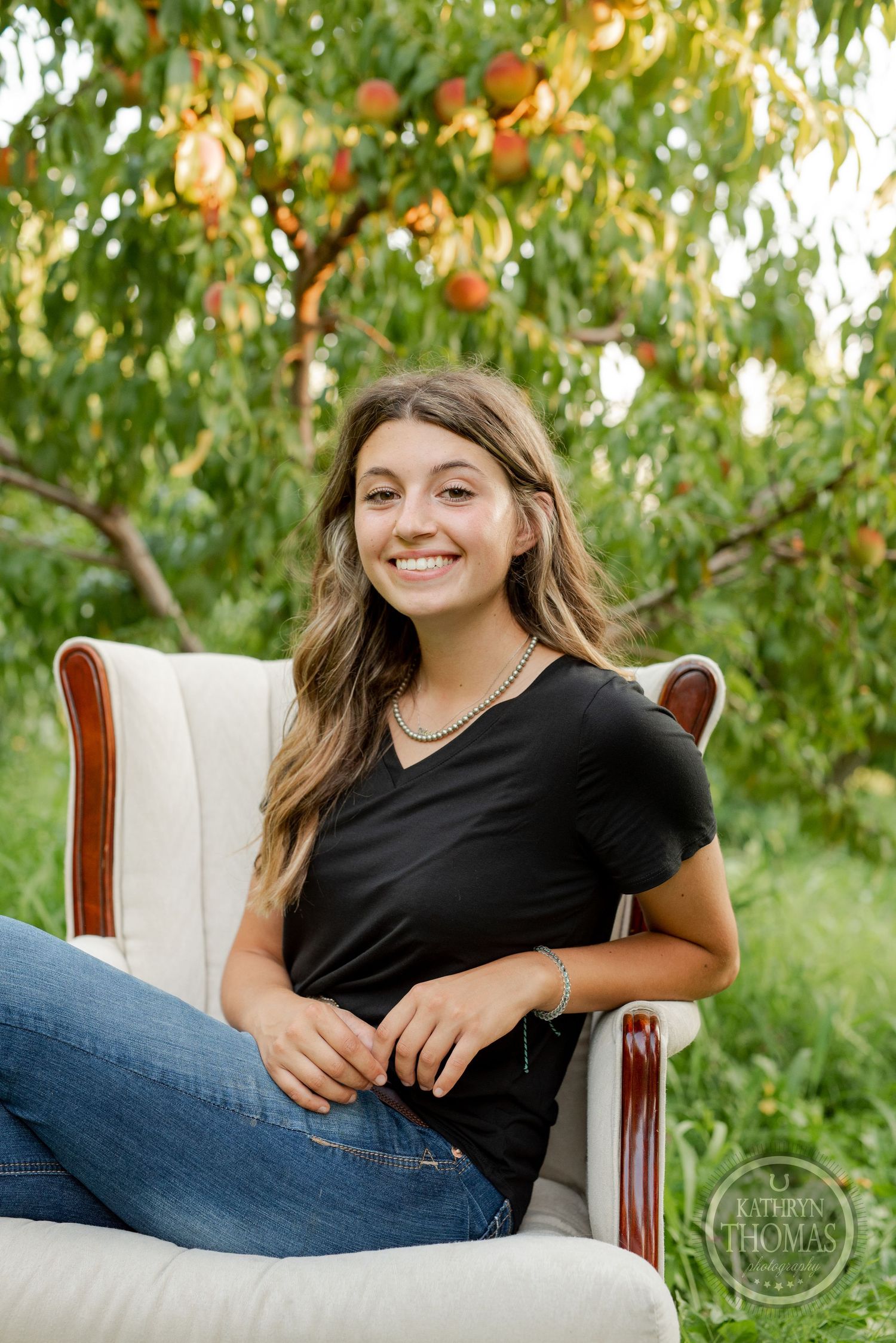 Casual portrait of someone relaxing in a white armchair against a peach tree backdrop in warm sunlight.