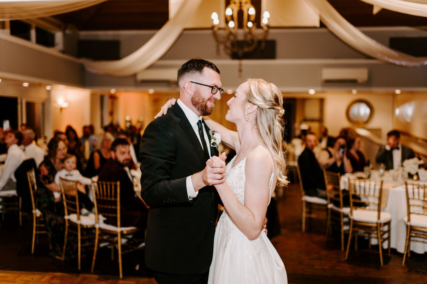 Couple dancing together at an elegant wedding reception with warm lighting and white decor.