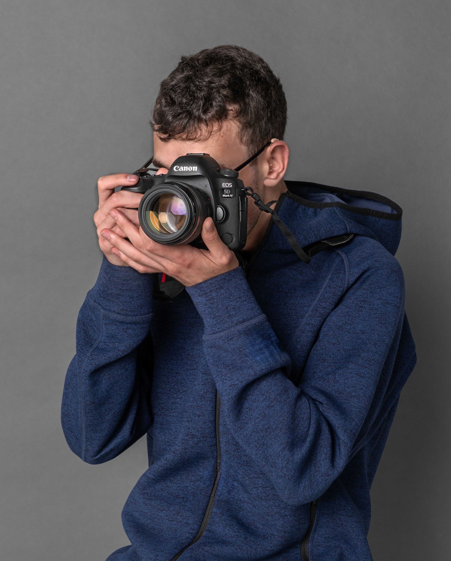Person in blue hoodie taking a photo with a professional DSLR camera against gray background.