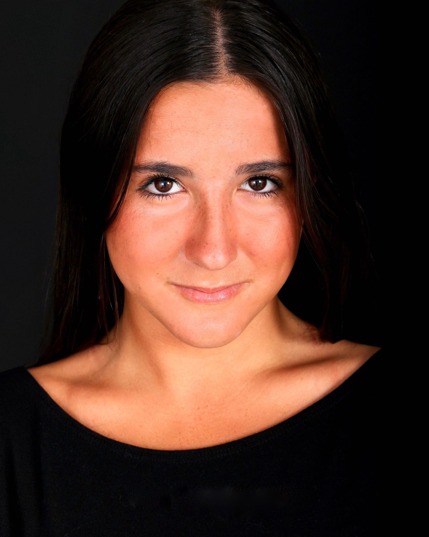 A dramatic headshot portrait of a young woman who has dark hair and dark eyes, set on a dark background.