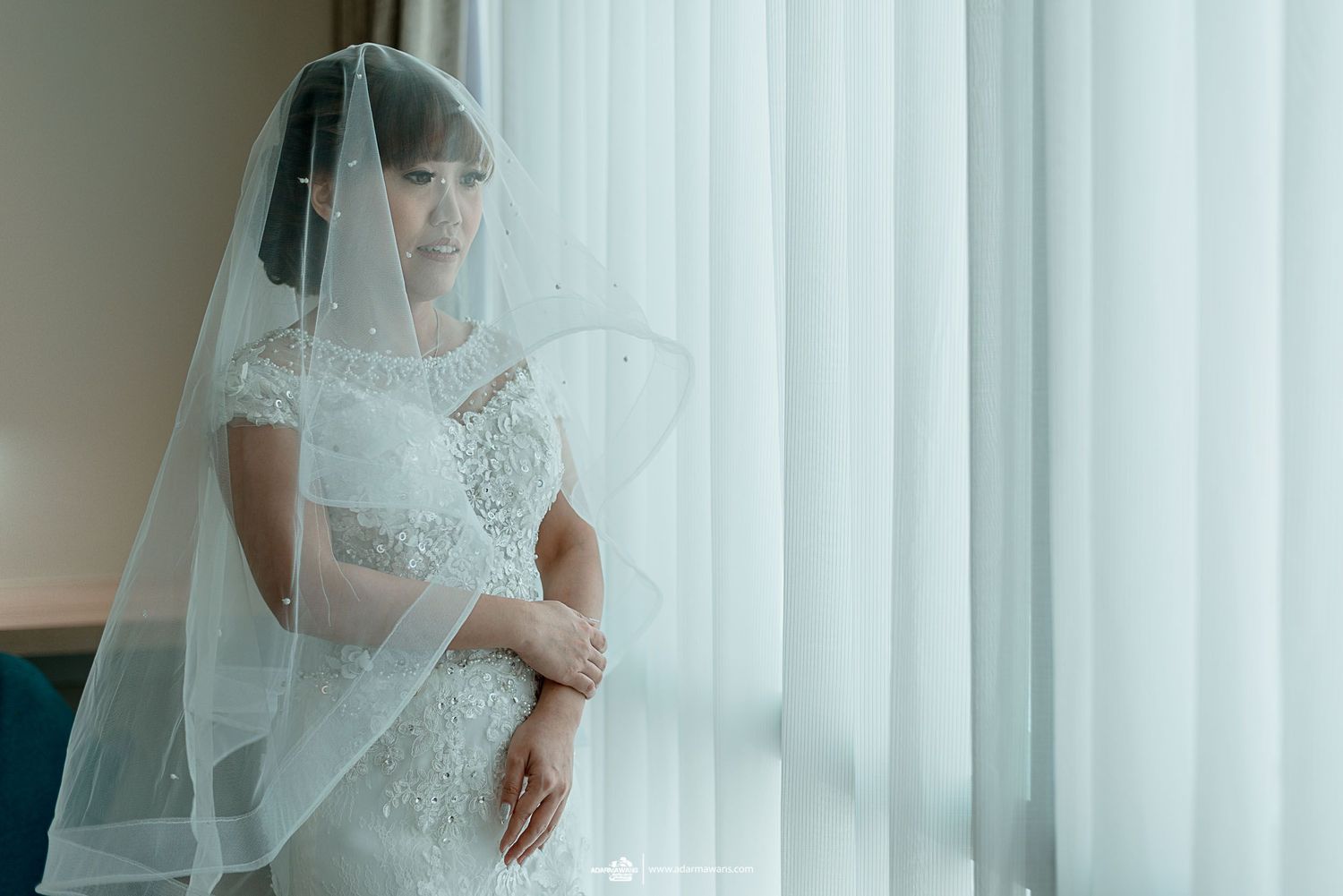 A bride in white lace gazes through sheer curtains in soft natural light.