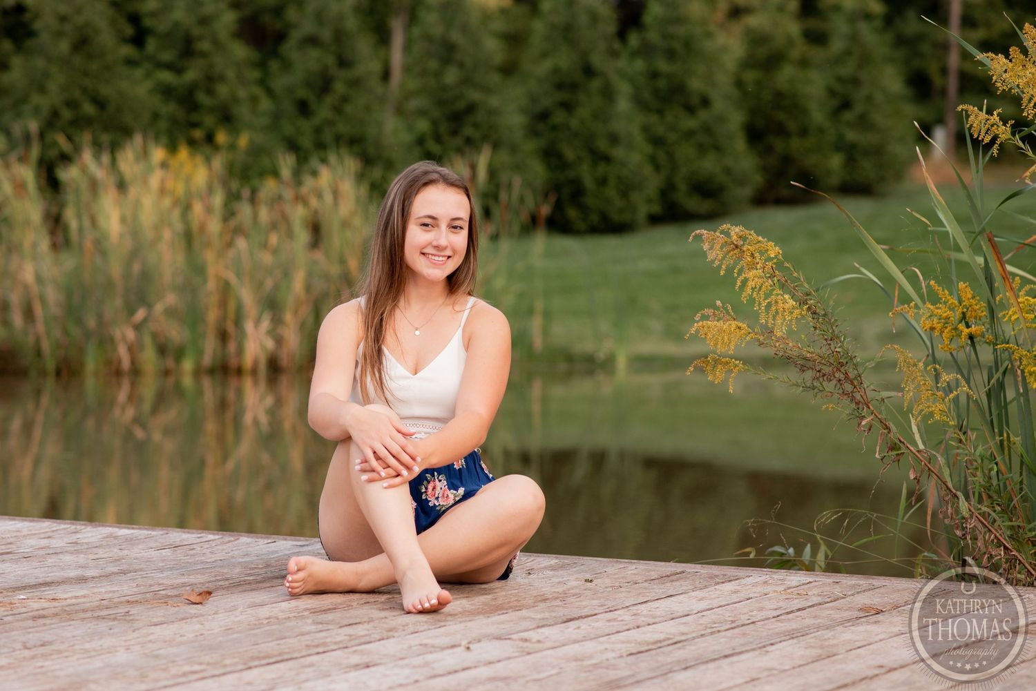 A casual outdoor portrait taken on a wooden dock with golden cattails and water in the background.
