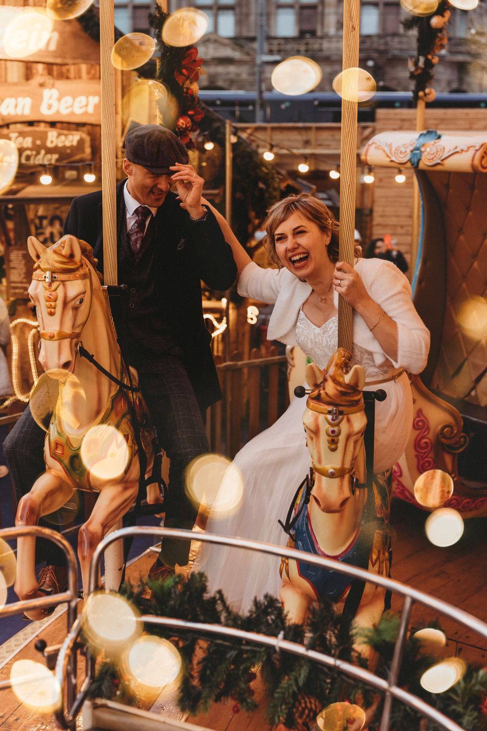 A couple enjoys a playful moment on carousel horses surrounded by twinkling lights.