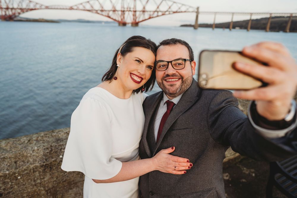 Couple taking selfie with iconic bridge in background during sunset.
