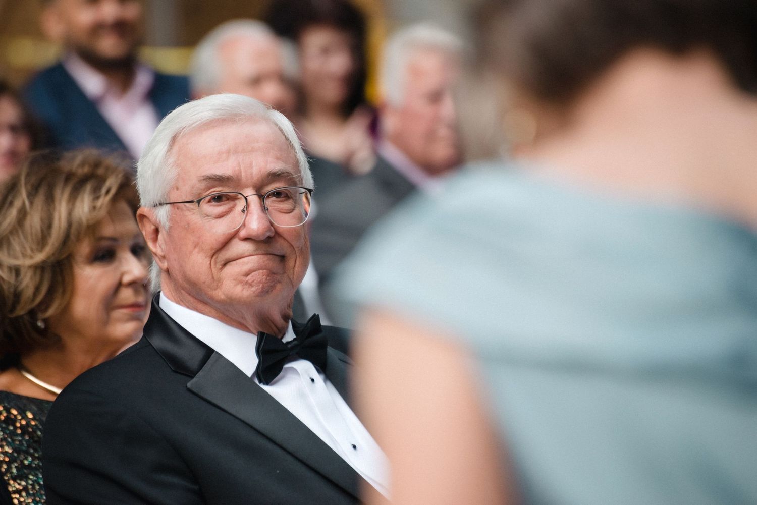 Father of the bride giving nod of approval at daughter's wedding with blurred person in foreground.