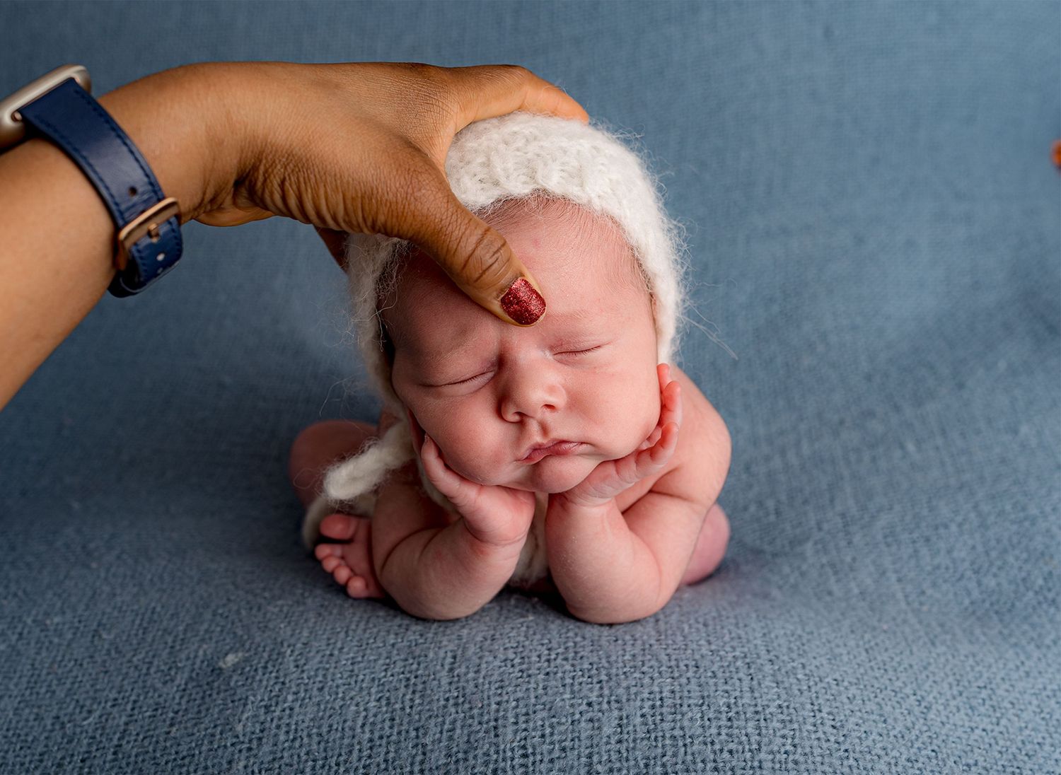 A froggy composition of a sweet sleeping newborn wearing white bonnet rests on blue backdrop.