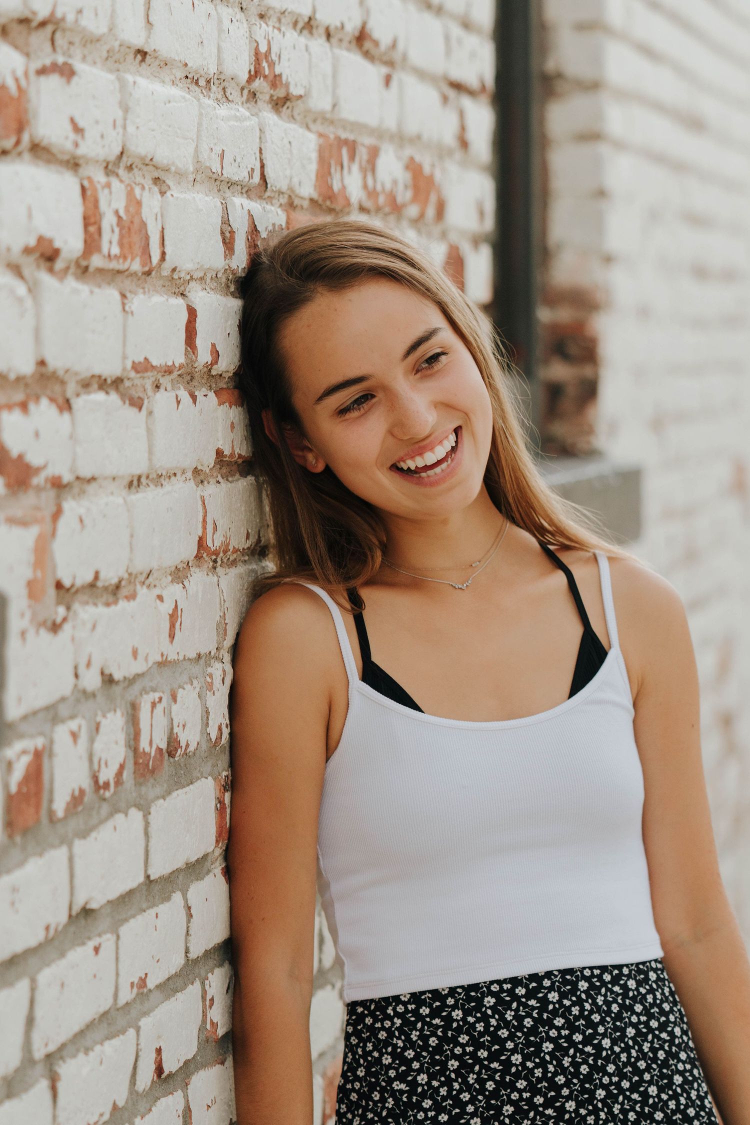 Portrait of a person in a white tank top and floral skirt smiling against a brick wall.
