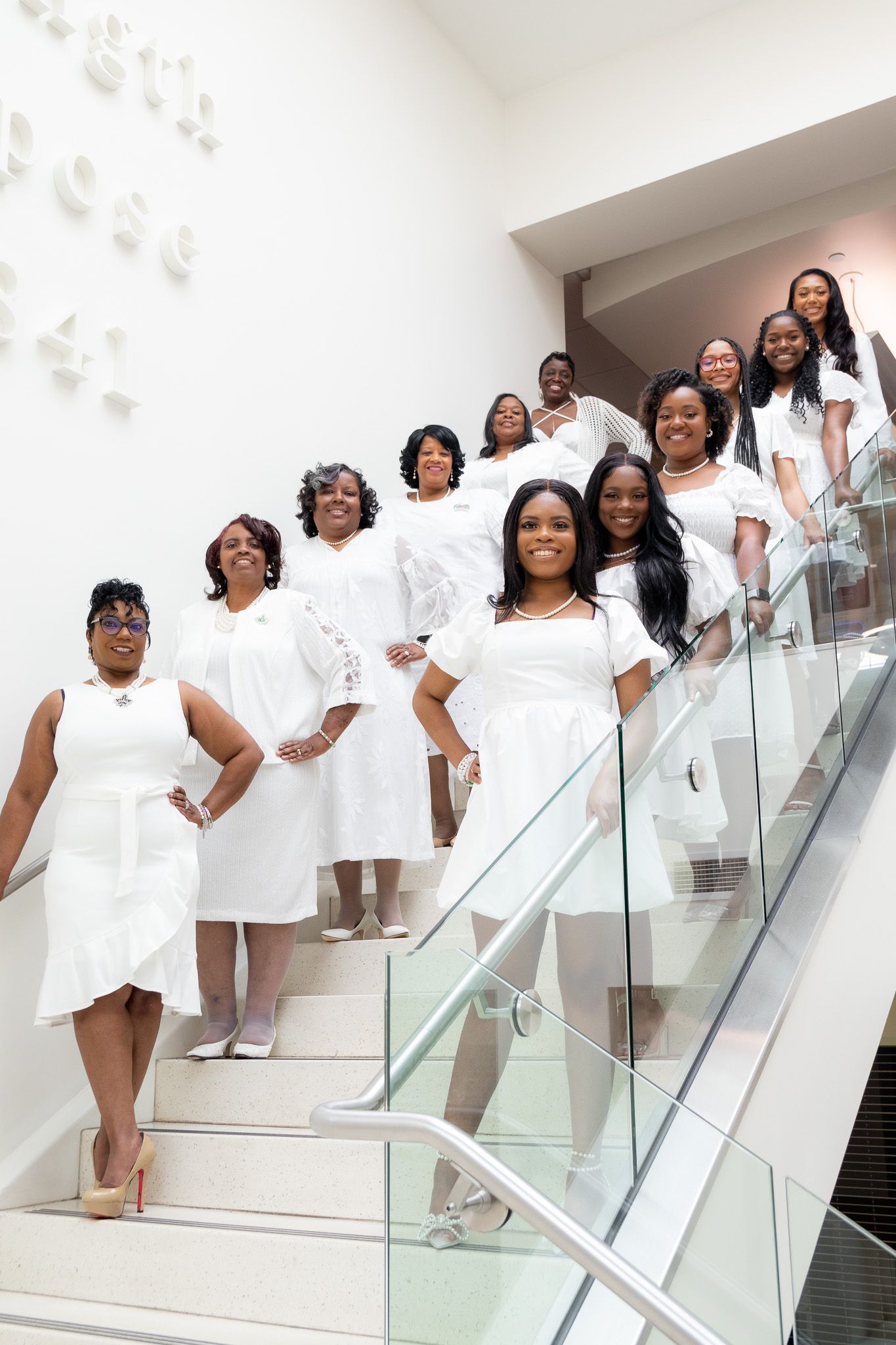 Group photo of women in white dresses posing on modern staircase.