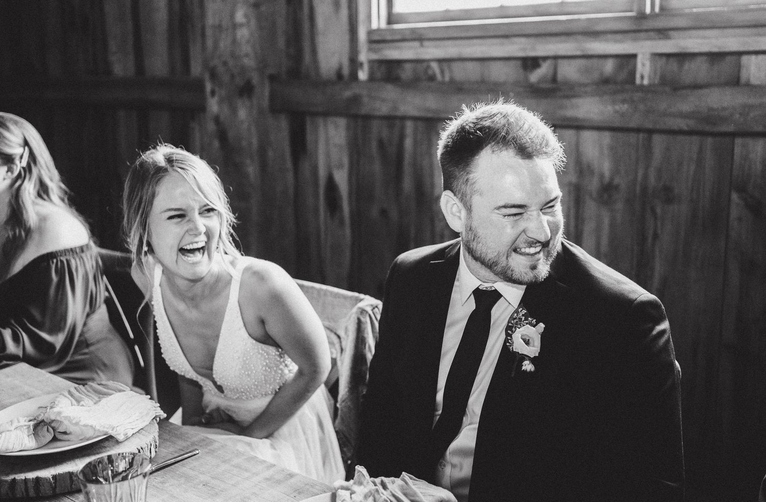A joyous black and white candid moment of laughter between wedding guests at a reception in a rustic barn setting.